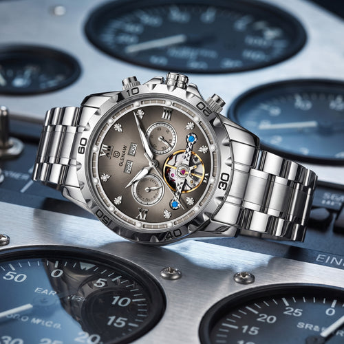 A luxury stainless steel wristwatch with a brown dial and a visible skeletonized automatic movement, displayed on a metallic panel with dials in the background.