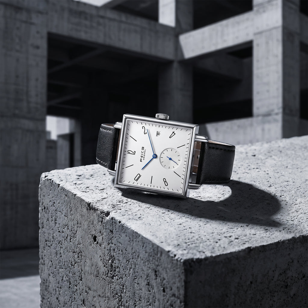 A rectangular silver-tone wristwatch with a white dial, black leather strap, and blue hands, displayed on a rough concrete block in an industrial concrete structure.