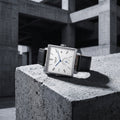 A rectangular silver-tone wristwatch with a white dial, black leather strap, and blue hands, displayed on a rough concrete block in an industrial concrete structure.