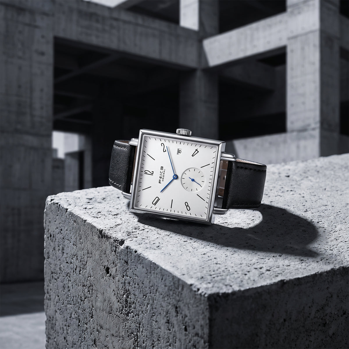 A rectangular silver-tone wristwatch with a white dial, black leather strap, and blue hands, displayed on a rough concrete block in an industrial concrete structure.
