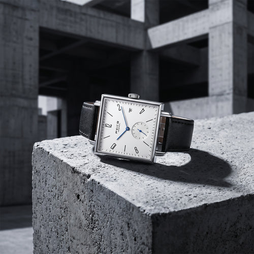 A rectangular silver-tone wristwatch with a white dial, black leather strap, and blue hands, displayed on a rough concrete block in an industrial concrete structure.