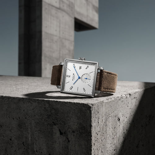 A rectangular silver-tone wristwatch with a white dial, black Arabic numerals, blue hands, and a brown suede strap, resting on a textured concrete ledge in an outdoor architectural setting.
