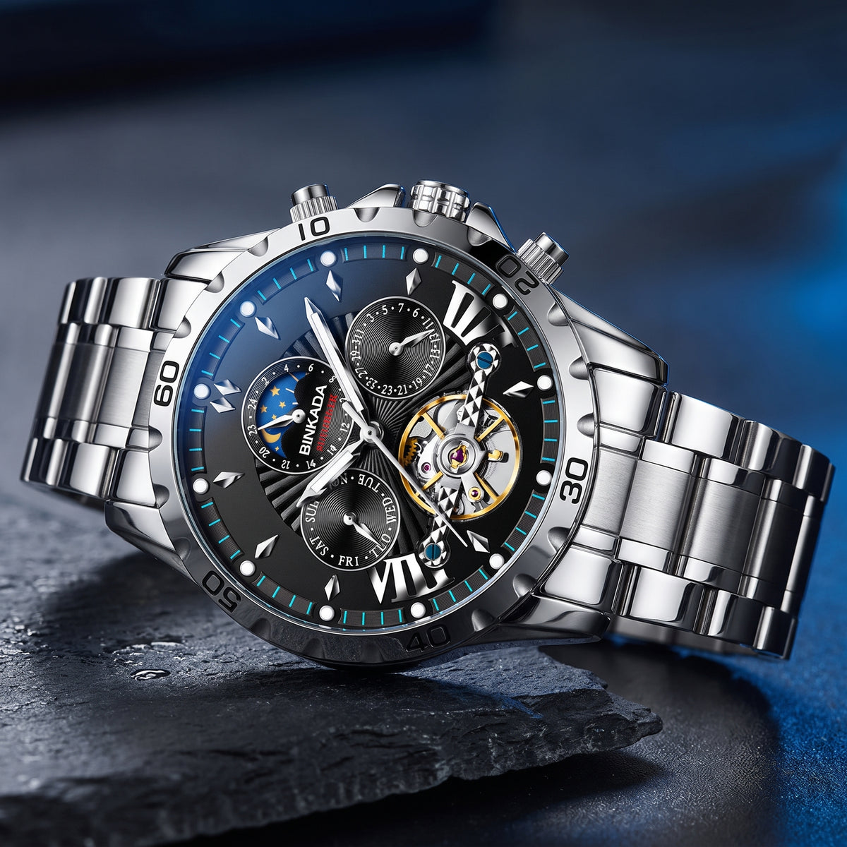 A stainless steel men's watch with a dark blue dial, featuring Roman numeral hour markers, subdials, a moon phase indicator, and a visible gold-tone skeletonized inner mechanism. The watch is resting on a dark slate surface with a blue gradient background.