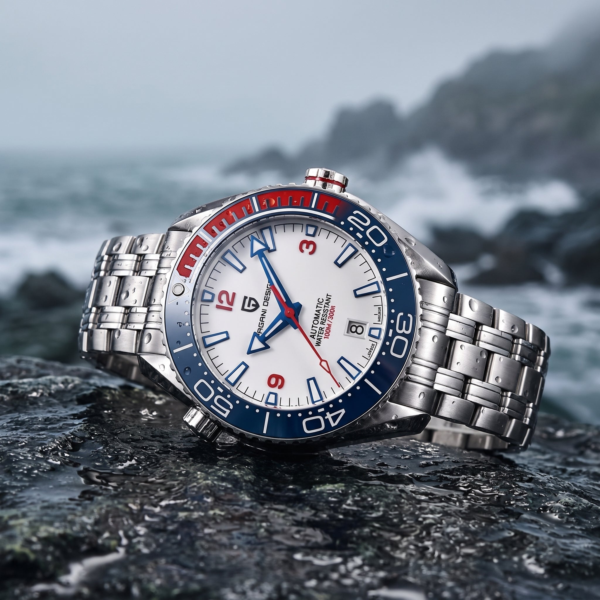 A stainless steel wristwatch with a silver metal link bracelet, featuring a white dial, a blue and red rotating bezel, and blue/red hands, photographed resting on dark rocks with a blurred ocean and rocky coastline in the background.