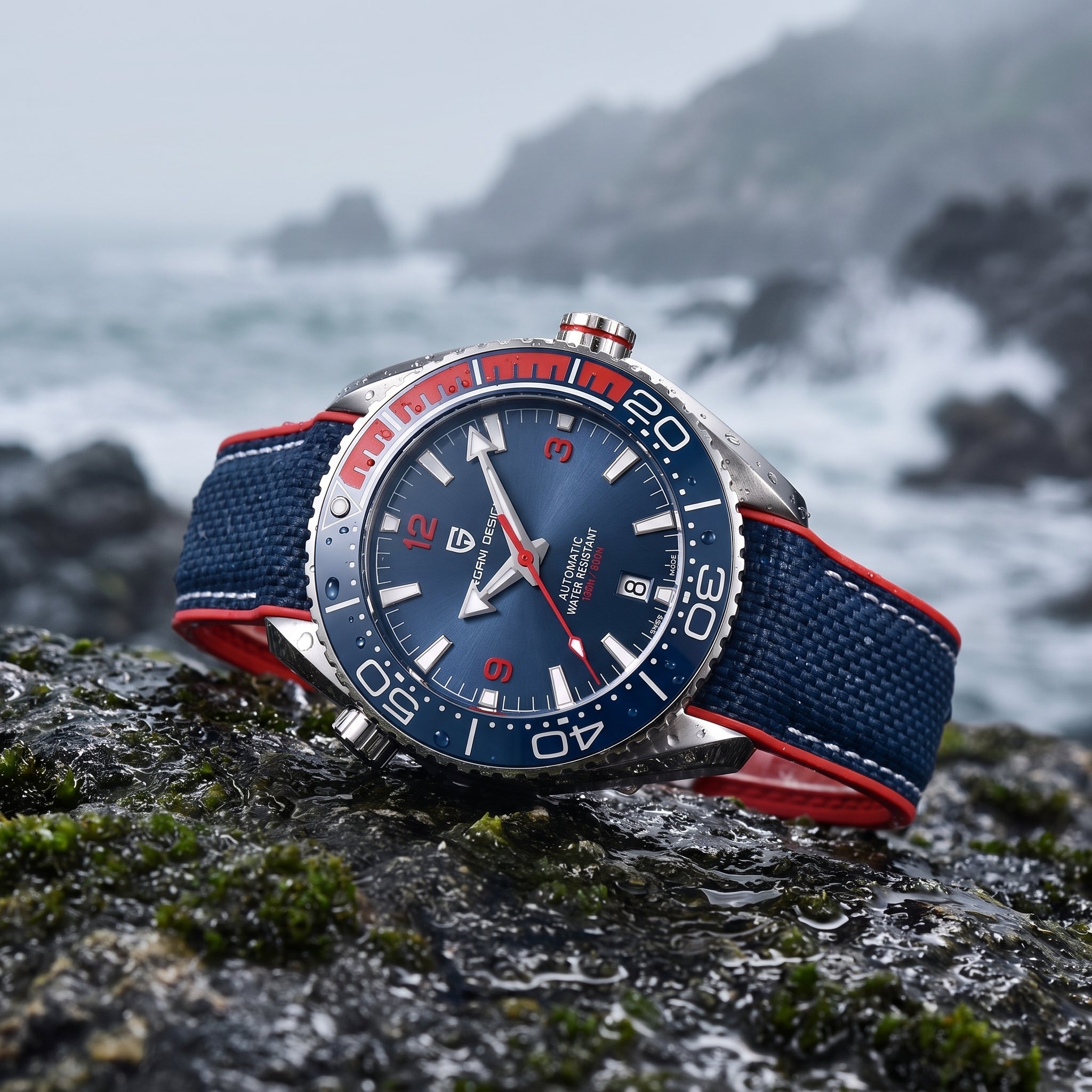 A wristwatch with a blue dial, silver-toned case, and blue-and-red fabric strap, photographed resting on a wet rock with a blurred coastal scene in the background.