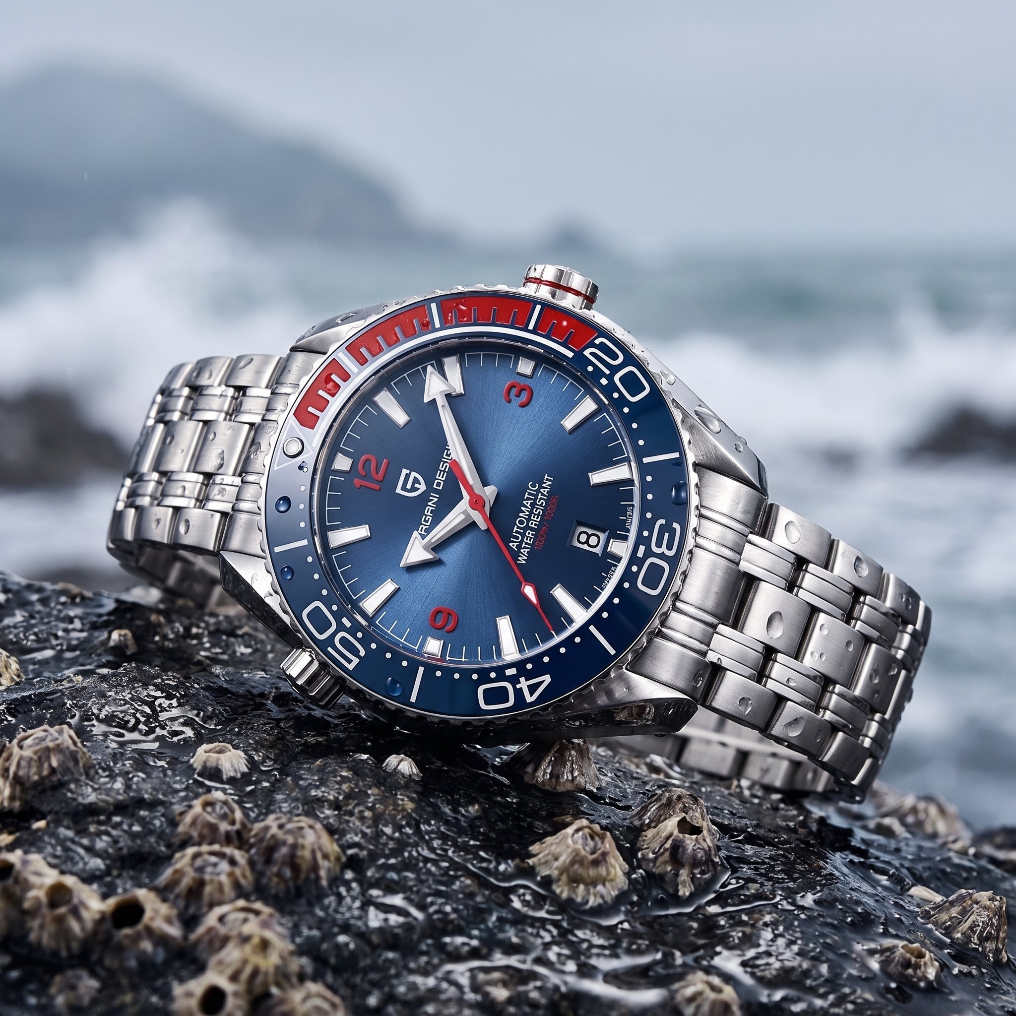 A stainless steel wristwatch with a blue dial and red and black bezel, resting on a wet rock with barnacles in the foreground and a blurred ocean background.