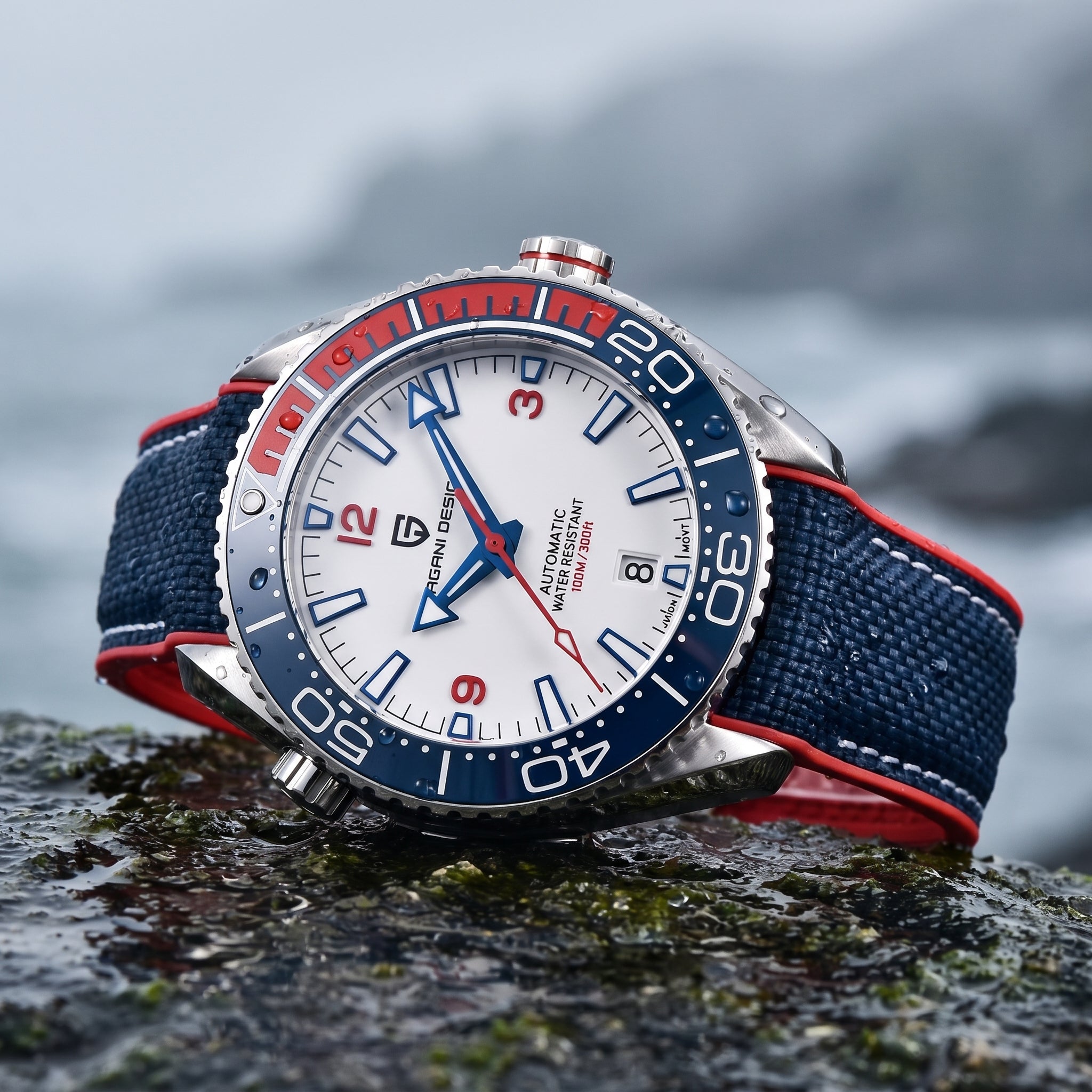 A wristwatch with a white dial, blue and red bezel, and navy blue fabric strap with red stitching, resting on a wet rock with a blurred coastal background.