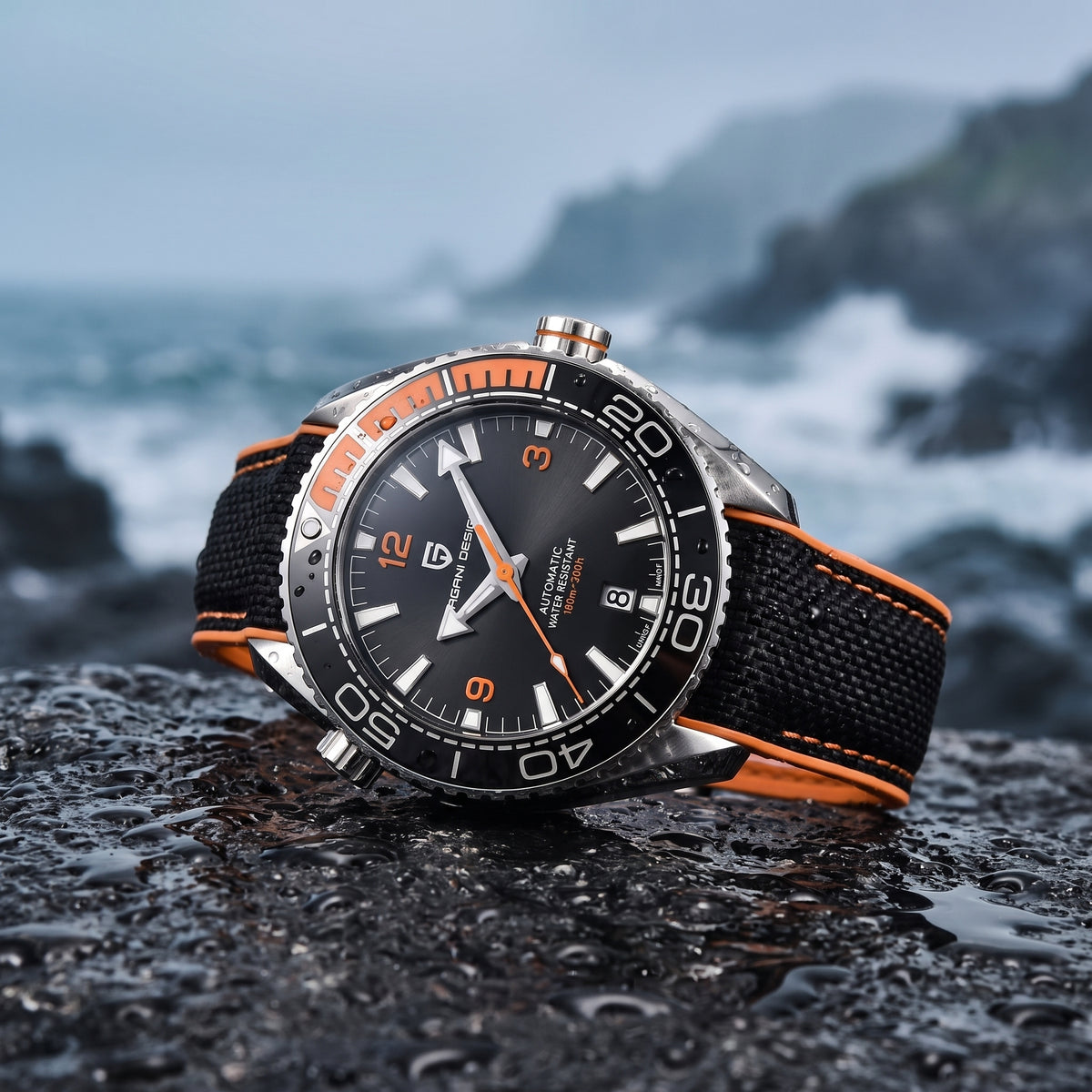 A wristwatch with a black dial, black and orange bezel, and black fabric strap with orange stitching, resting on wet dark rocks with a blurred ocean and rocky coastline in the background.