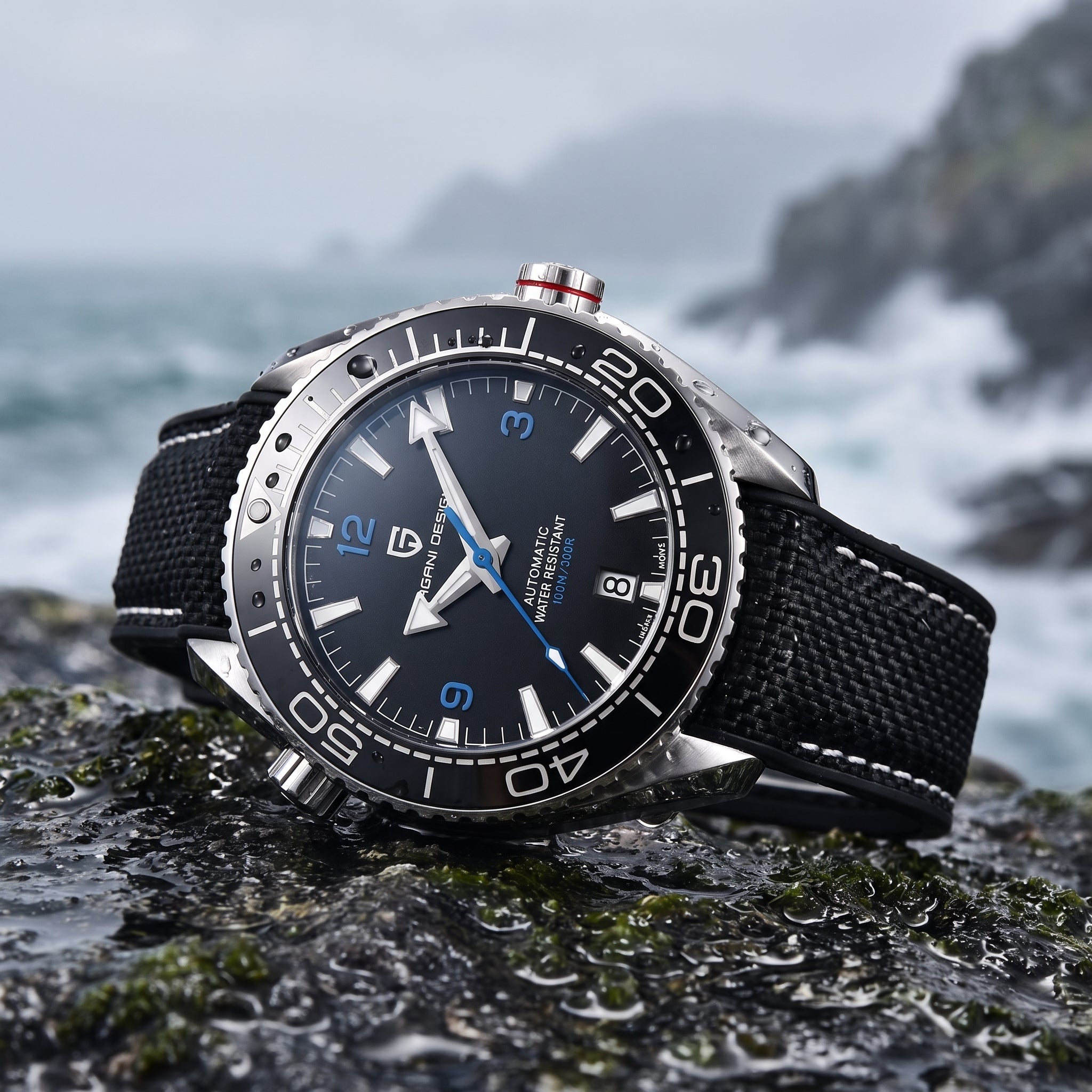 A wristwatch with a black dial, silver-toned case, and black woven strap, photographed resting on a wet rock with a blurred ocean and rocky coastline in the background.