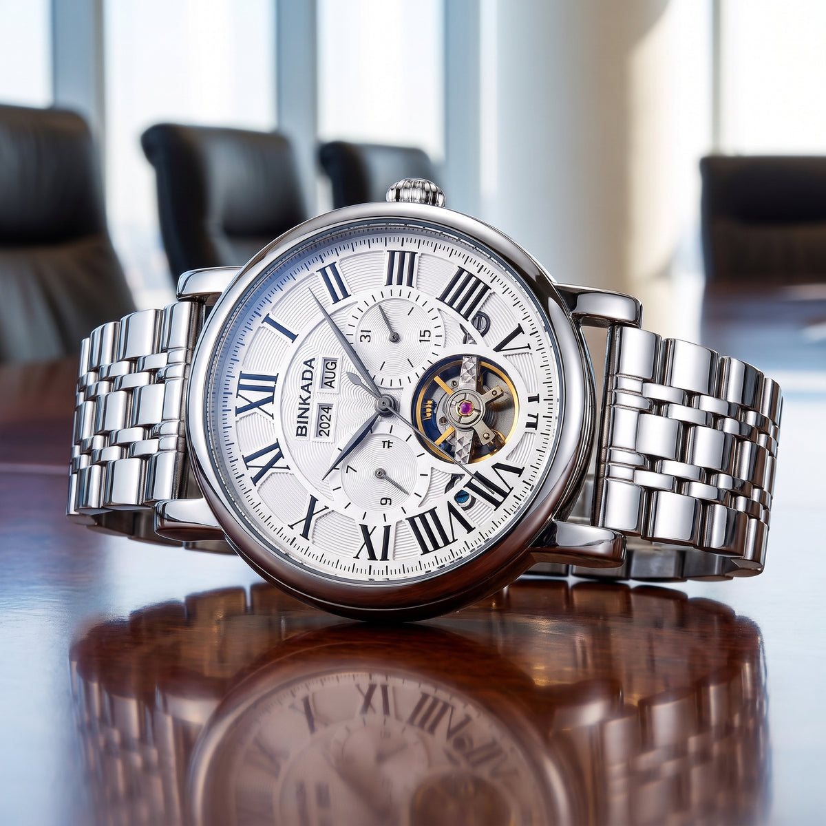 A silver-tone BINKADA wristwatch with a stainless steel link bracelet, featuring a white dial with Roman numerals, a visible tourbillon, and a date window, displayed on a reflective wooden table.